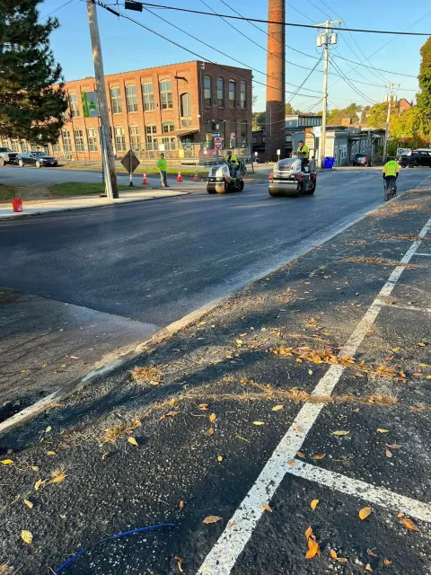 Street with freshly paved asphalt, vehicles, workers, and a brick building.