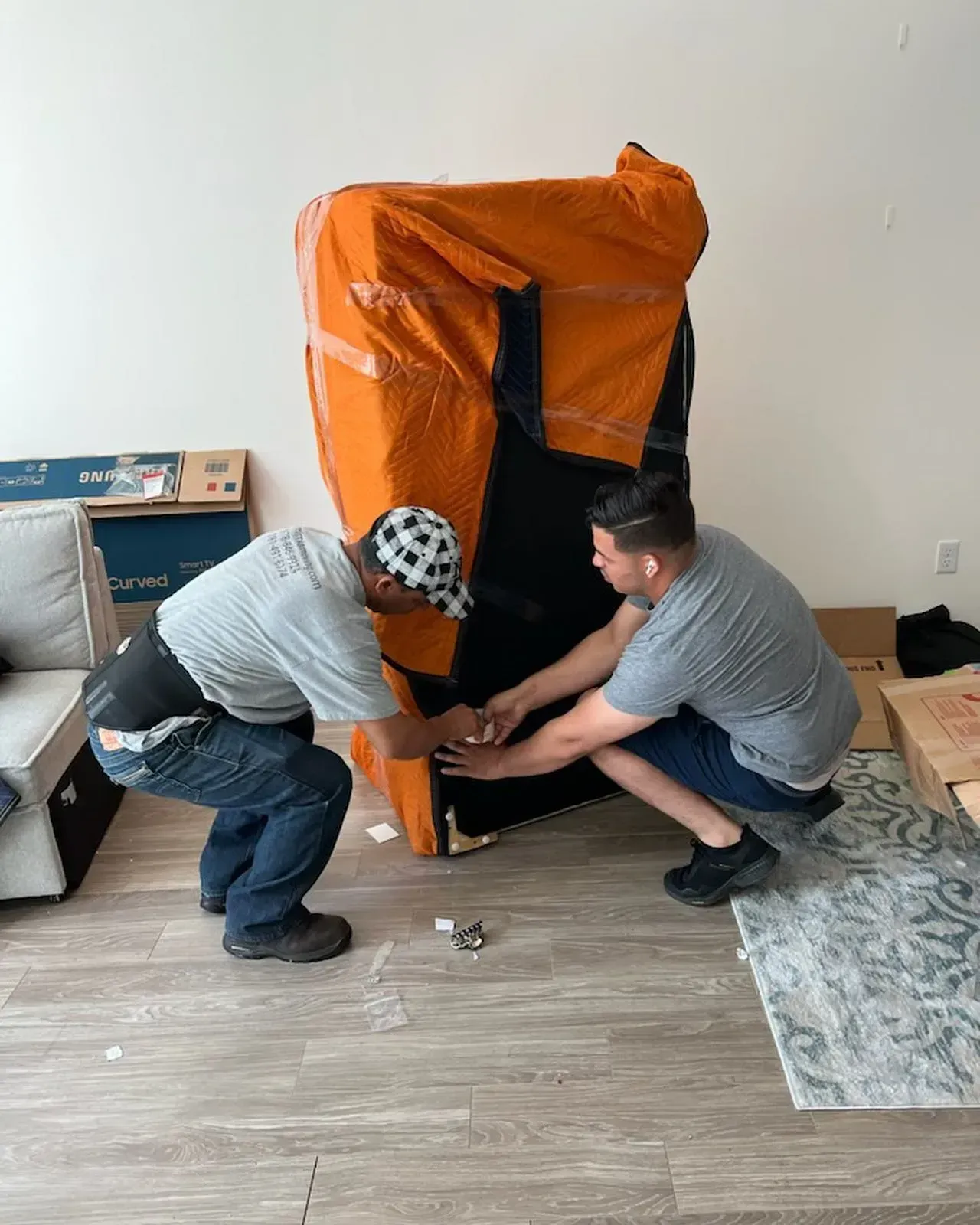 Two people lifting a large, wrapped object inside a room. The object is orange and black.