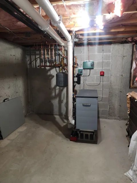 Basement with a grey boiler, pipes, and a fire alarm on a concrete wall.