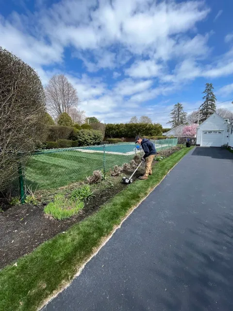 A man using a weed trimmer to trim grass along a garden bed next to a paved driveway under a partly cloudy sky.