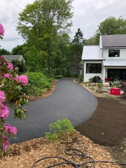 Asphalt driveway curves toward a white house with landscaping. Pink flowers and mulch border.