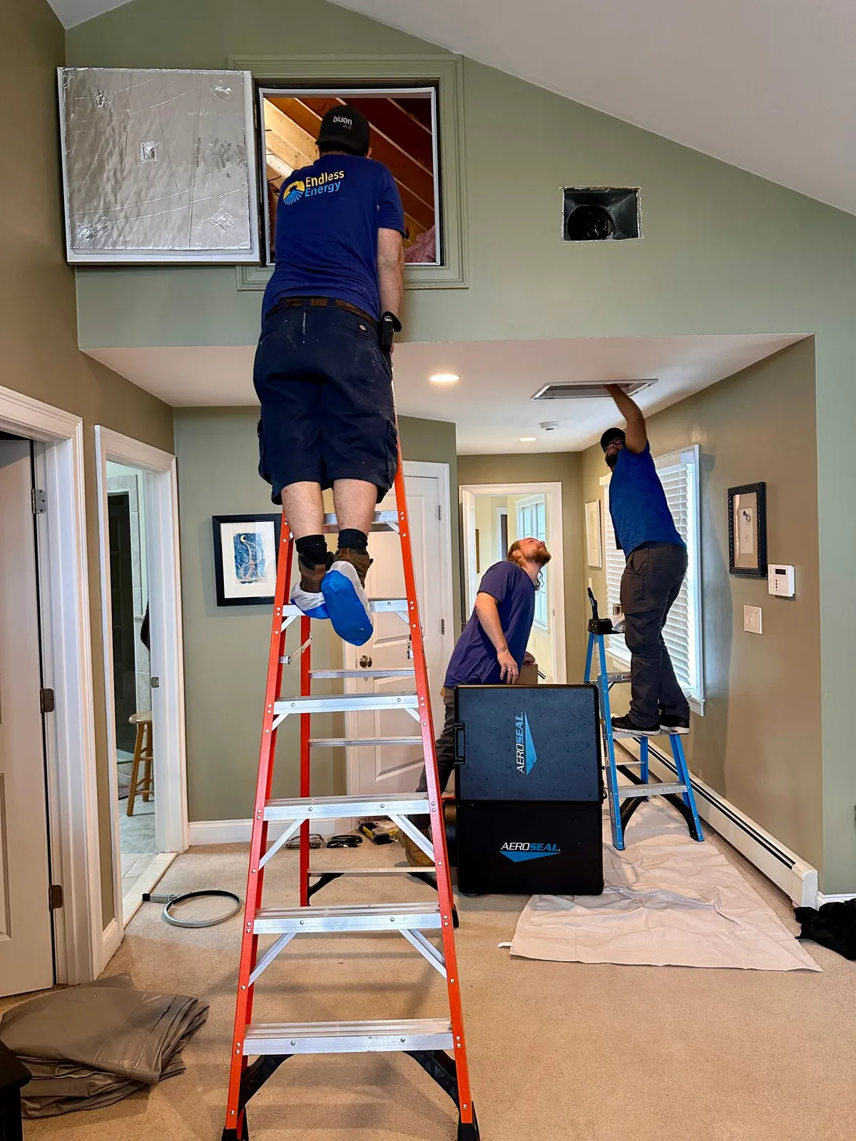 Three workers on ladders install or repair ductwork and ventilation in a home, using tools and equipment, with protective sheets covering the floor.
