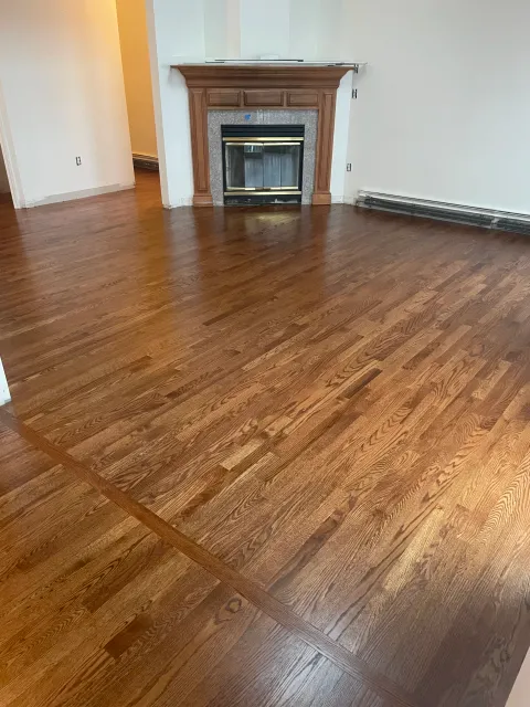 A room with hardwood flooring and a brick fireplace with wooden trim. The walls are painted white, and there are no furnishings.