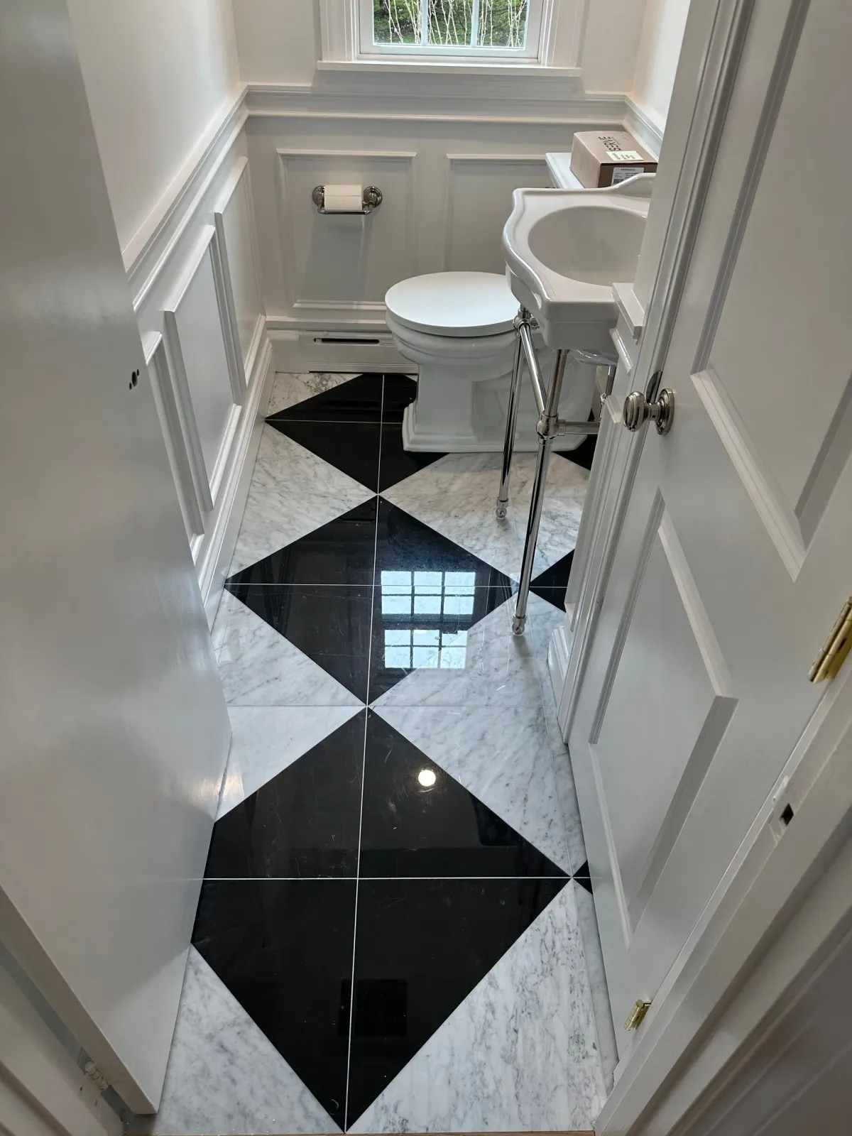 Narrow bathroom with black-and-white diamond tile floor, toilet, sink, and window above.