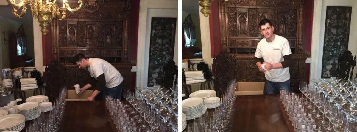 A person arranges white tableware and many wine glasses on a long wooden table in a formal, dimly lit dining room.