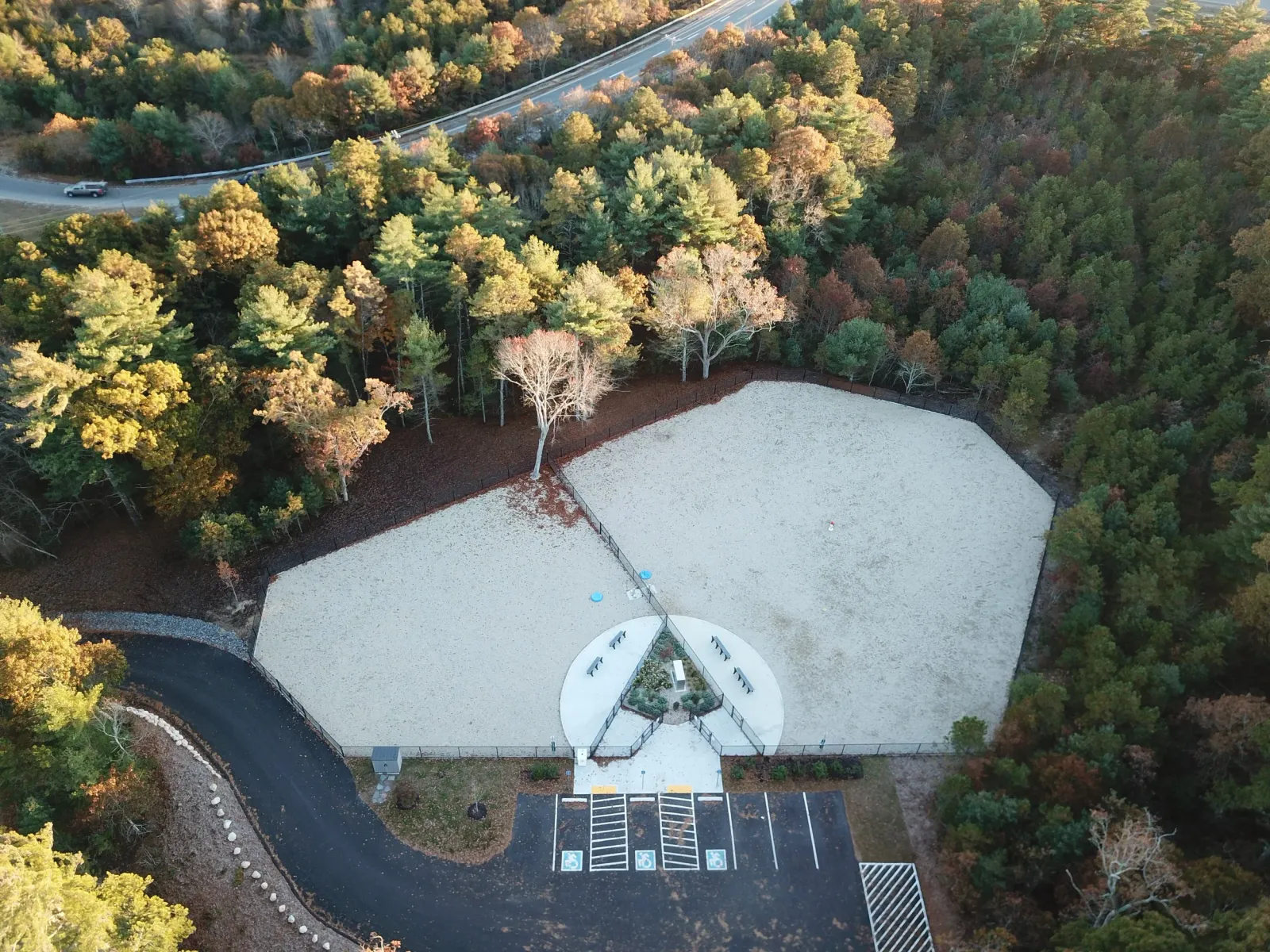 A bird's-eye view of a small, octagonal outdoor ice skating rink surrounded by trees with colorful fall foliage, with a parking lot and a winding road nearby.