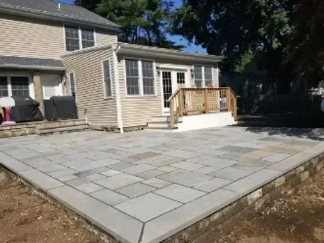 Backyard patio with gray pavers and a raised wall, connecting to a house with a sunroom.