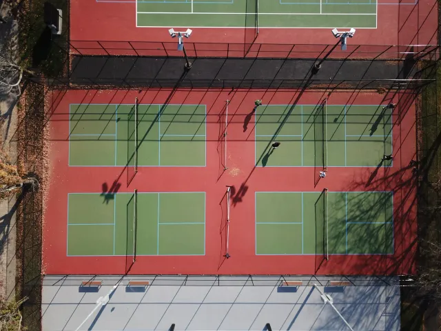 An aerial view of a tennis court with four courts, separated by white lines and equipped with nets. The courts are surrounded by black fencing and tall lights. Four people are playing, with their shadows cast on the surface.