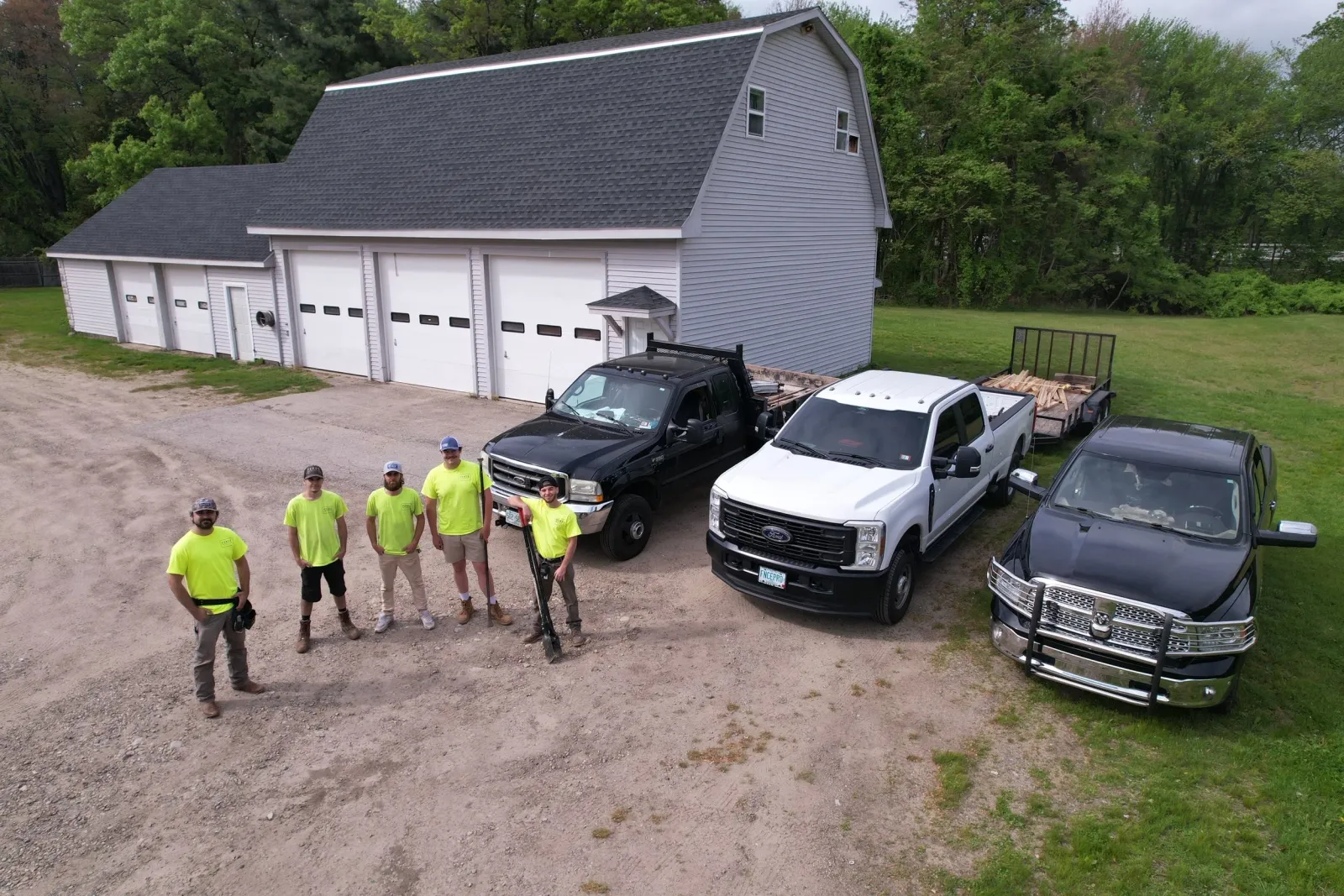 Group of six men in bright yellow shirts standing in front of parked trucks near a large gray barn and green trees.