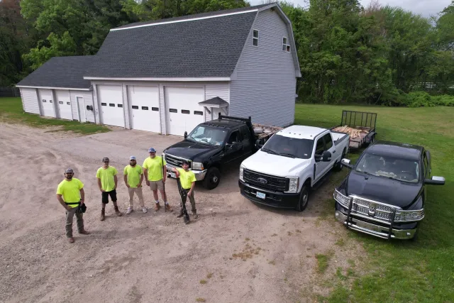 Group of six men in bright yellow shirts standing in front of parked trucks near a large gray barn and green trees.