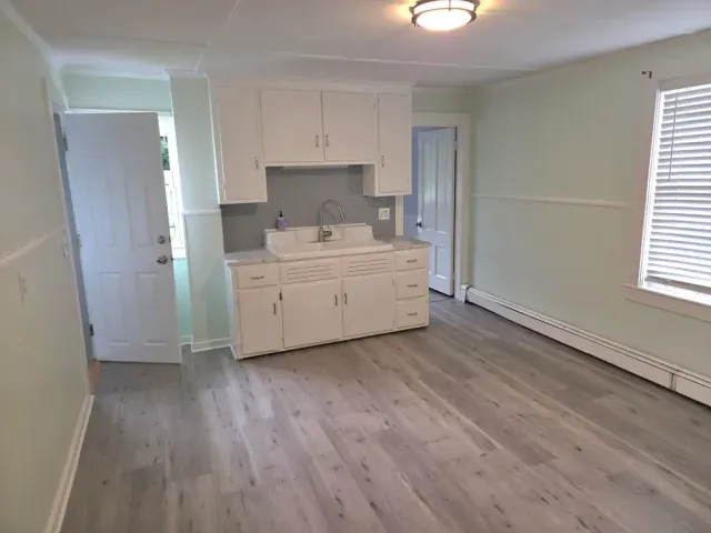 Empty kitchen and living area with white cabinets, gray flooring, and a window with blinds.