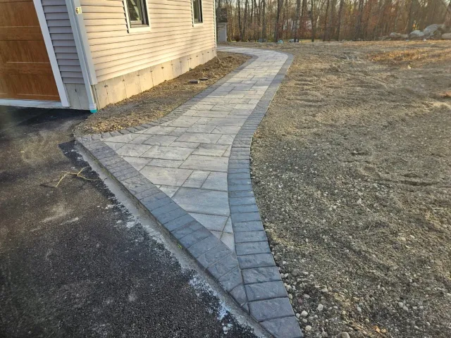 A paved walkway curves from a driveway towards a house. Gravel surrounds the path.