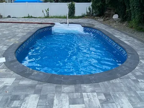 An oval shaped swimming pool is surrounded by a brick patio.