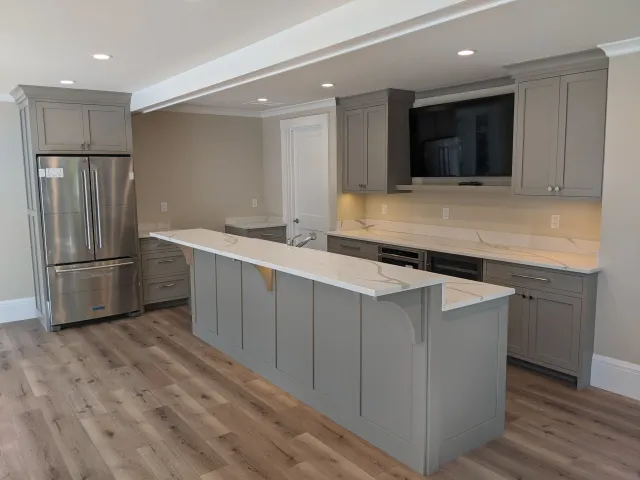 Modern kitchen with gray cabinets, white marble countertops, stainless steel refrigerator, hardwood flooring, and a wall-mounted television.