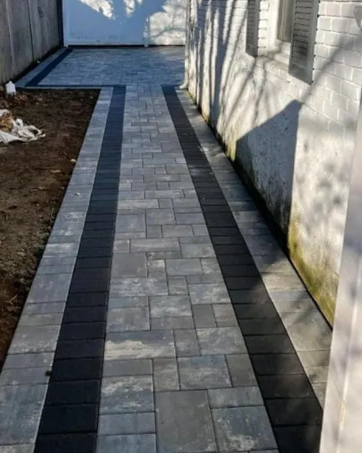 A newly installed stone paver walkway with a grey-toned center and dark border runs alongside a white brick house wall.