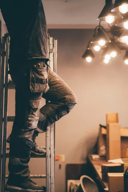 A person wearing work clothes on a ladder with boxes and tools in a workshop or storage area, under hanging ceiling lights.