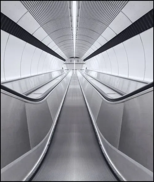 A photograph depicting an escalator seamlessly integrated within a brightly lit tunnel. The tunnel is illuminated by intense, white lighting, emphasizing a blend of modern and industrial design elements. The metallic sheen of the escalator stands out starkly against the bright backdrop, leading towards a vanishing point in the distance.