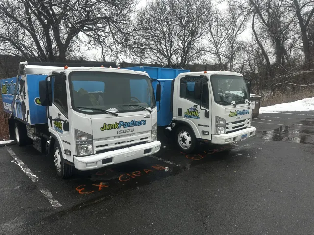 Two white Junk Panthers Isuzu trucks with blue dump bodies parked in a lot after a snowfall