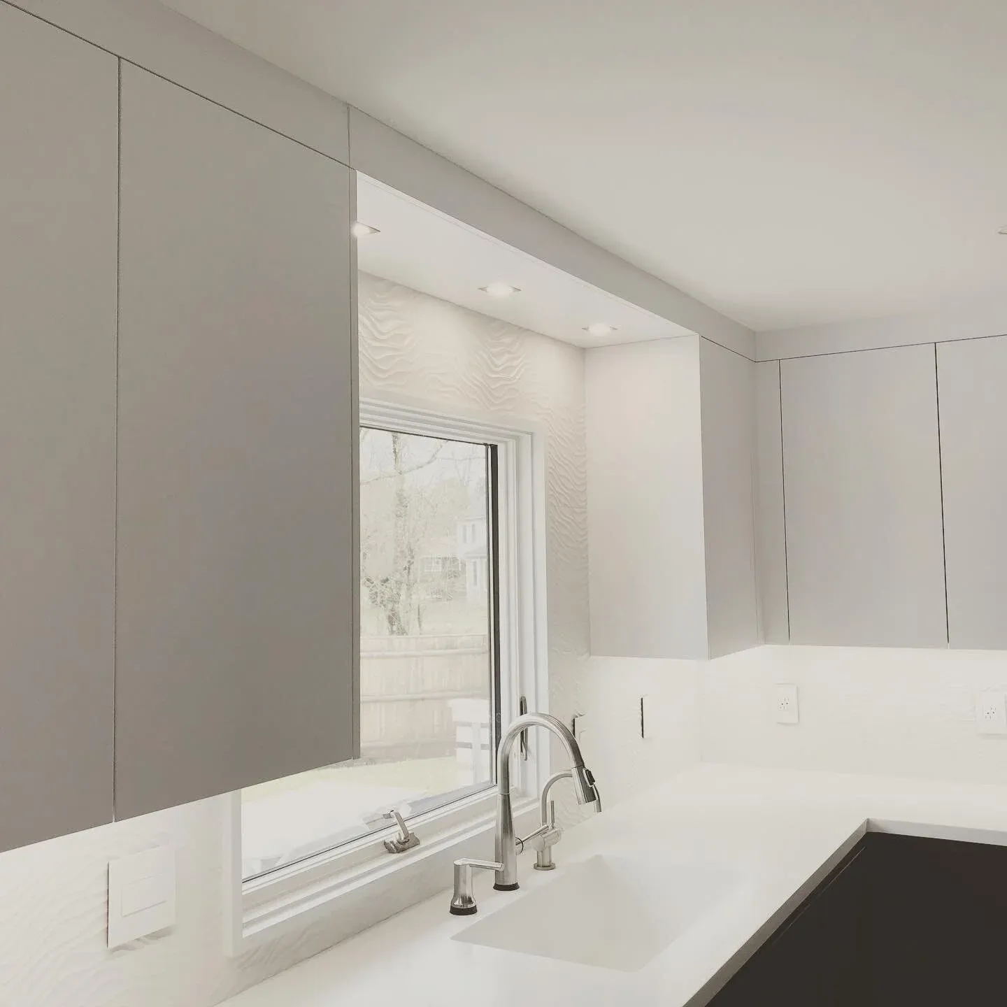 Modern kitchen interior with gray cabinets, a large window, and a white countertop featuring a built-in sink and faucet. Recessed lighting illuminates the space.