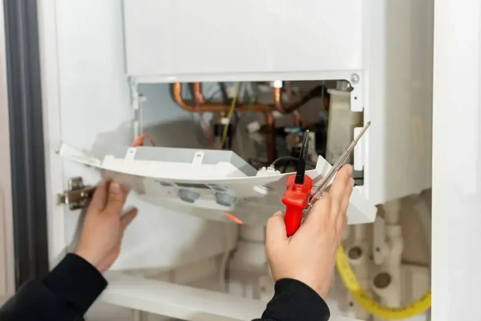 Technician's hands open a panel of a white appliance, exposing copper pipes and wiring inside and tools nearby.
