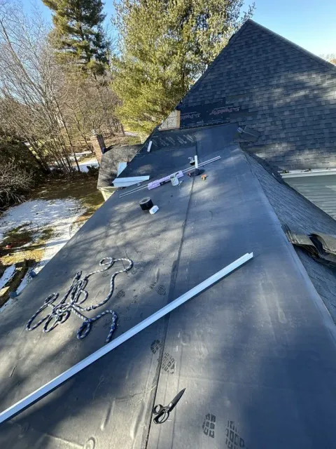Roof under repair with roofing materials and tools on a house beside trees and snow.