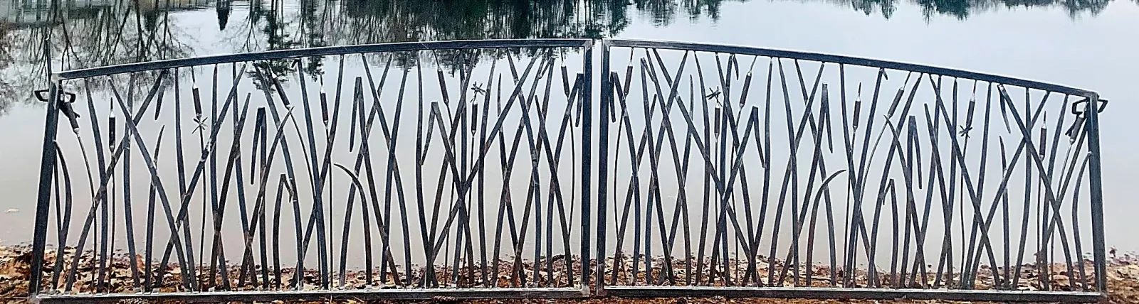 A black metal fence with a decorative, uneven pattern, situated in front of a calm body of water with trees reflected on its surface.