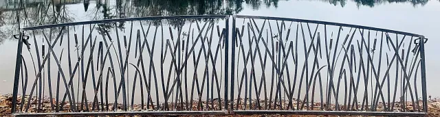 A black metal fence with a decorative, uneven pattern, situated in front of a calm body of water with trees reflected on its surface.
