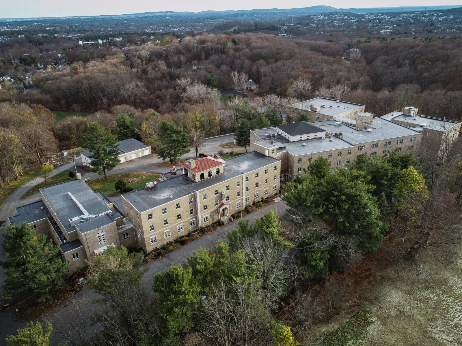 Aerial view of a large, rectangular stone building surrounded by trees, with a winding road and scattered smaller structures nearby; rolling hills and a suburban landscape are visible in the distance.