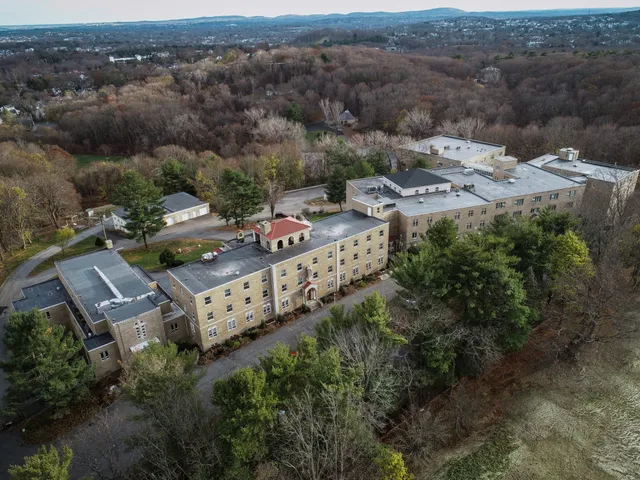 Aerial view of a large, rectangular stone building surrounded by trees, with a winding road and scattered smaller structures nearby; rolling hills and a suburban landscape are visible in the distance.