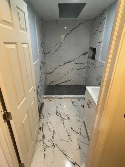 Bathroom with marble-like tiles on walls and floor, a walk-in shower area with a dark gray floor, a recessed niche for toiletries, and a white vanity with a sink on the right.