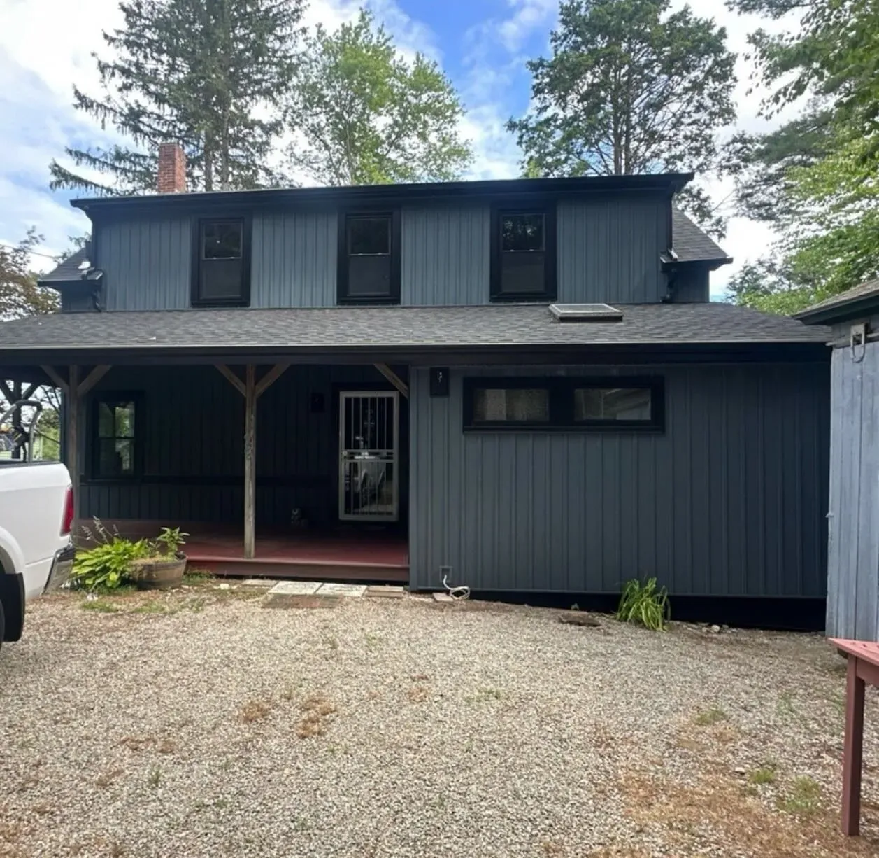 A dark gray, two-story house with a covered porch and gravel driveway under a bright blue sky with scattered clouds.