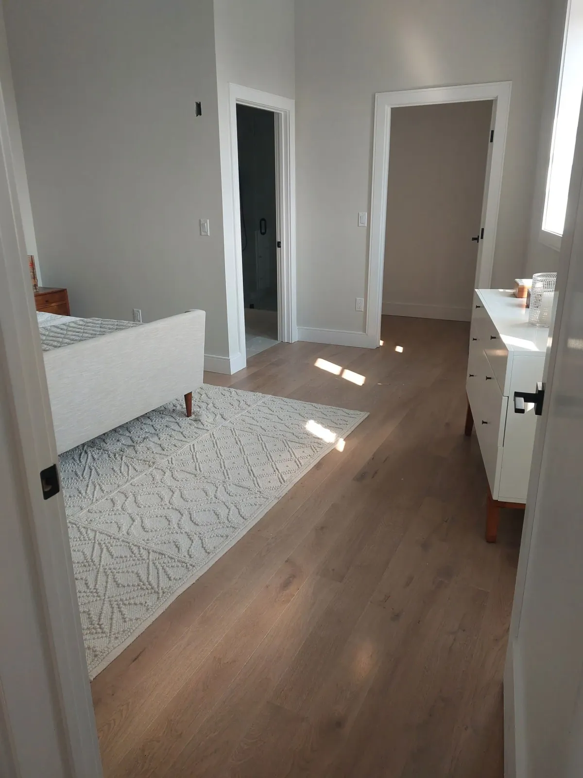 Bedroom with light wood floor, white dresser, and a fluffy rug. Doors and windows.