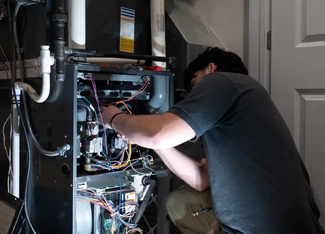 A person is repairing or inspecting the wiring inside an open furnace unit in a utility room.