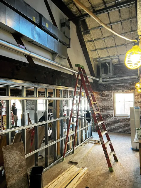 Interior construction scene with ladder, ductwork, exposed metal framing, and brick walls