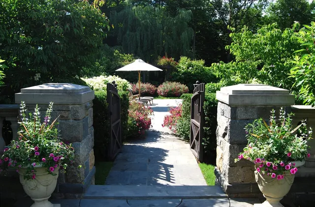 A serene garden path flanked by flowering plants leads to a shaded table with an umbrella, surrounded by lush greenery and stone accents.