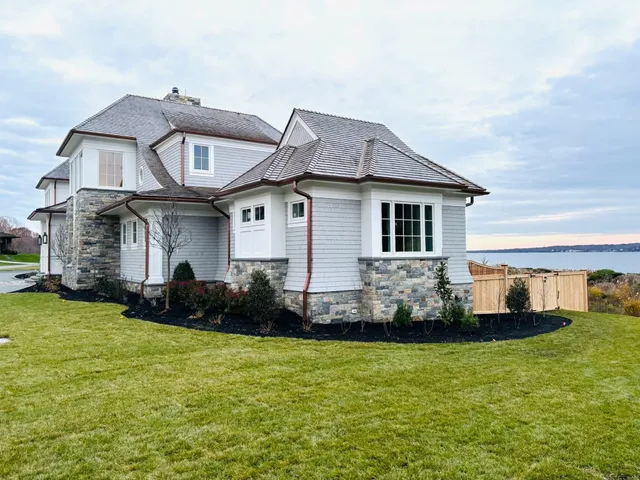 White and stone coastal home with green lawn and water view