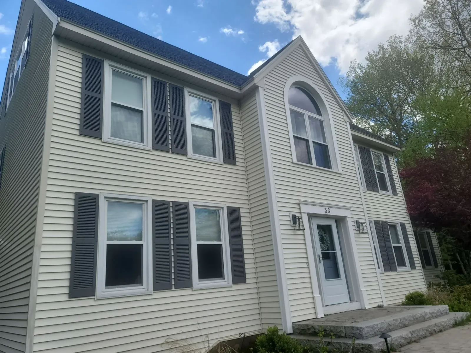 Two-story gray house with white trim, blue shutters, and a front porch under a partly cloudy sky