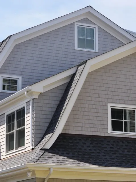 The side of a home with gray shingle siding, a dark roof, and multiple gambrel-style dormer windows.