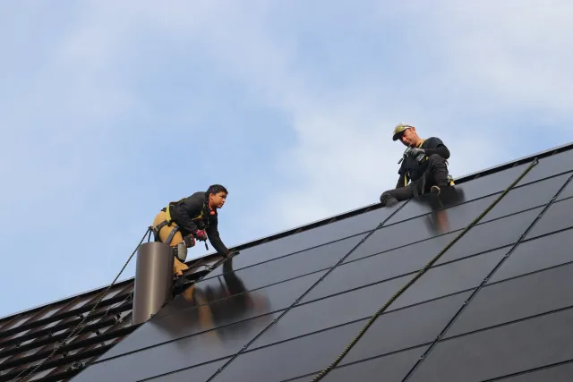 Two workers on a sloped metal roof, wearing safety gear, working on installing or maintaining the roofing.