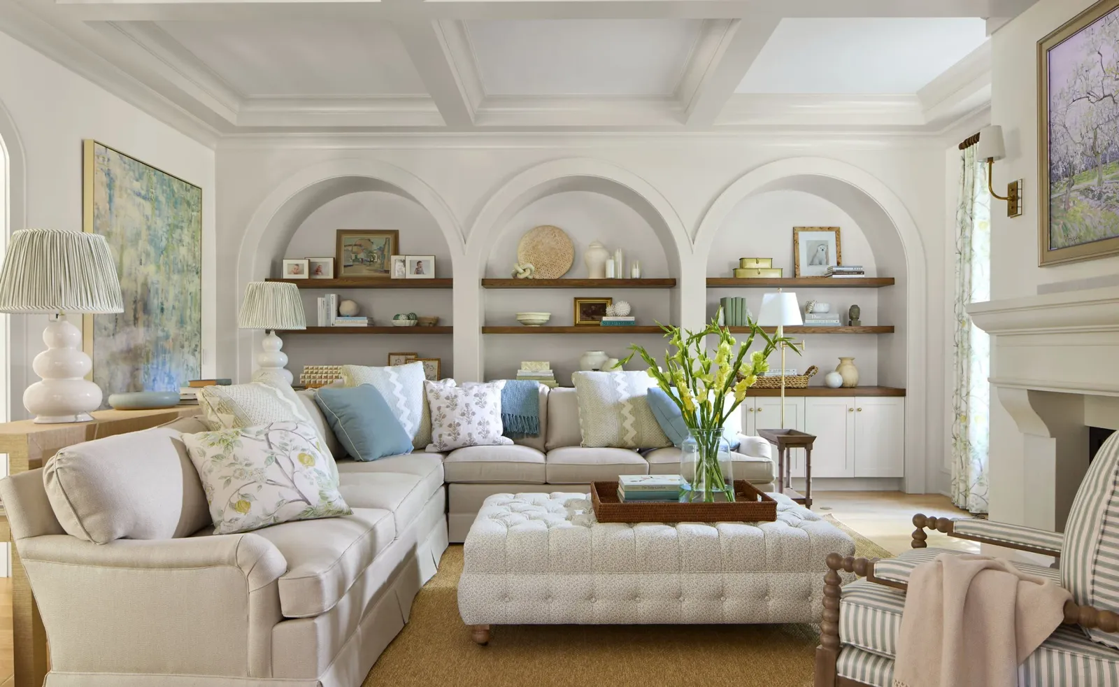 Elegant living room with a beige sectional sofa, decorative pillows, and a tufted ottoman. Shelves with artwork and decor items are set against arched recesses. A vase with flowers and table lamps complement the serene atmosphere. Paintings and soft