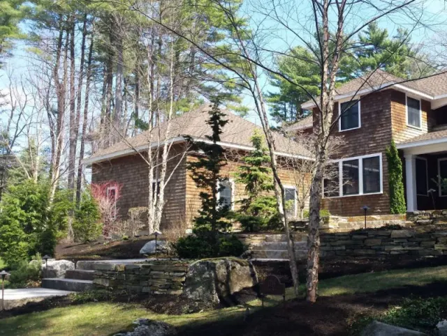 Two-story wooden house surrounded by tall pine trees and landscaped stone retaining walls in a forested area.