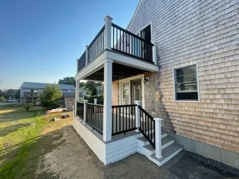 Side view of a house with wooden shake siding, featuring a two-level deck with black railings and stairs, on a sunny day.