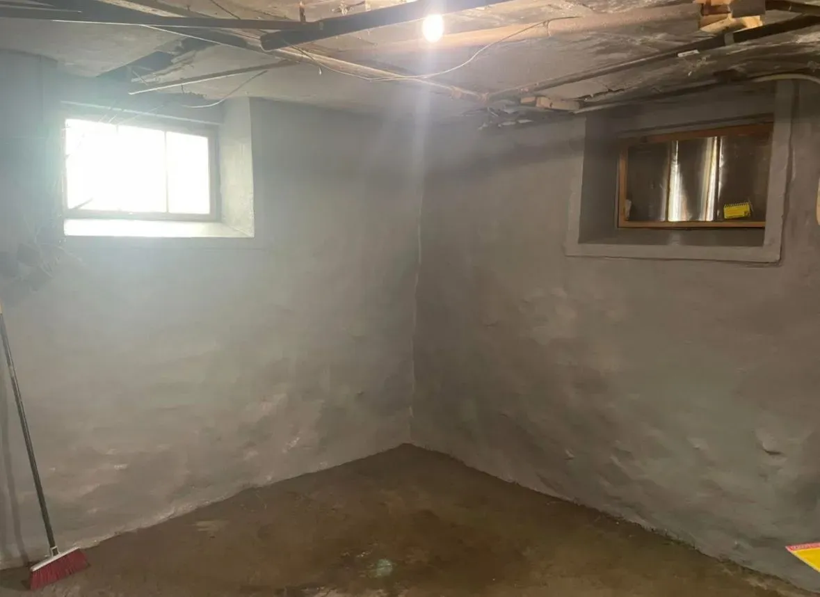 Bare basement room with gray walls, exposed ceiling beams, two small windows, and a concrete floor.