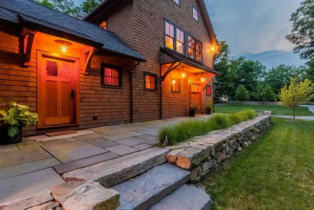 Exterior of a rustic wooden house with warm lighting, stone pathway, and landscaped yard with small tree and grass.