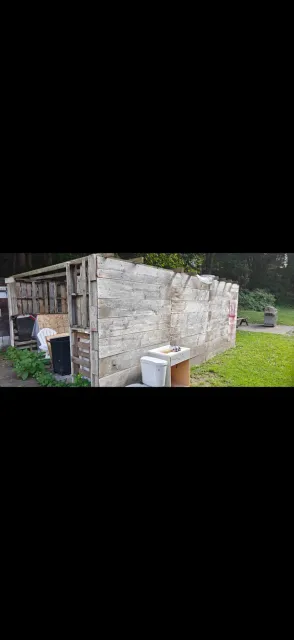 Small stone shed with a wooden door in a grassy yard, with buckets and a barrel beside it.