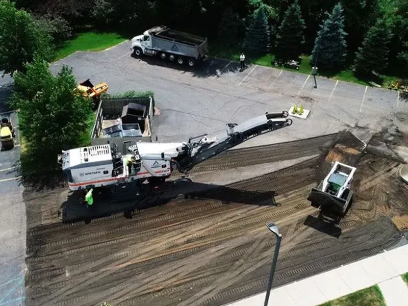 Asphalt milling in progress on a parking lot. Equipment includes a milling machine, trucks, and a skid steer.