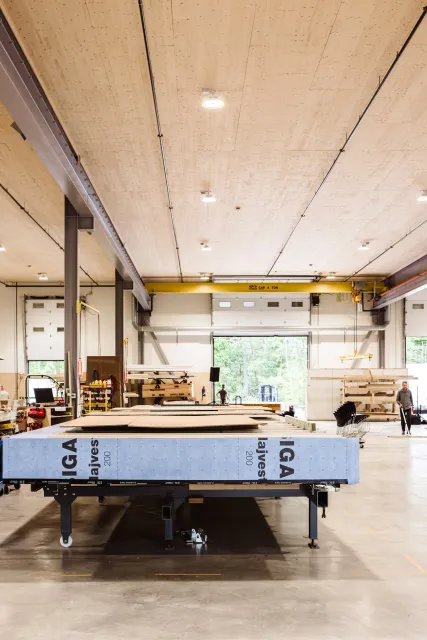 Inside a woodworking shop with large work tables, storage shelves, and workers. The ceiling has a yellow overhead crane labeled "CAP. 4 TON" and large garage doors open to outside greenery.