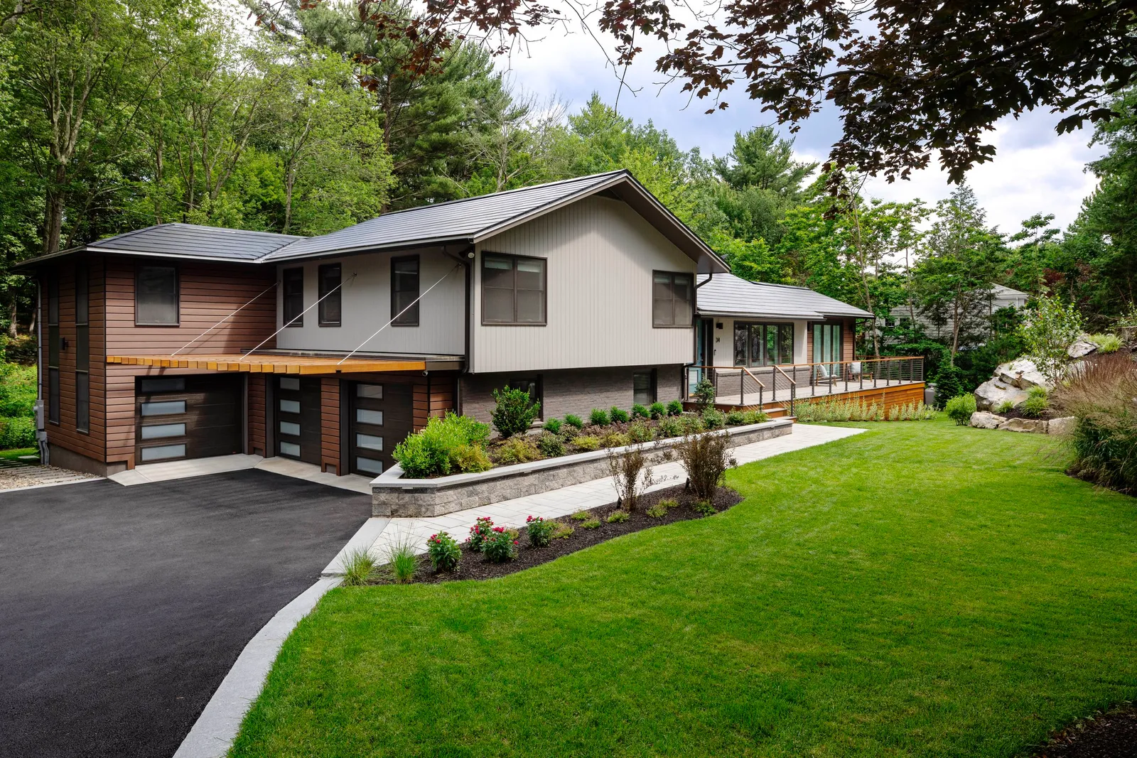 Modern house with a black driveway, landscaped yard, and large deck, surrounded by green trees.