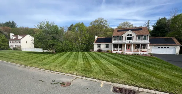 A large, two-story house with beige siding and a brown shingle roof, featuring solar panels, a front porch with white columns, and a well-maintained lawn with alternating light and dark green stripes.