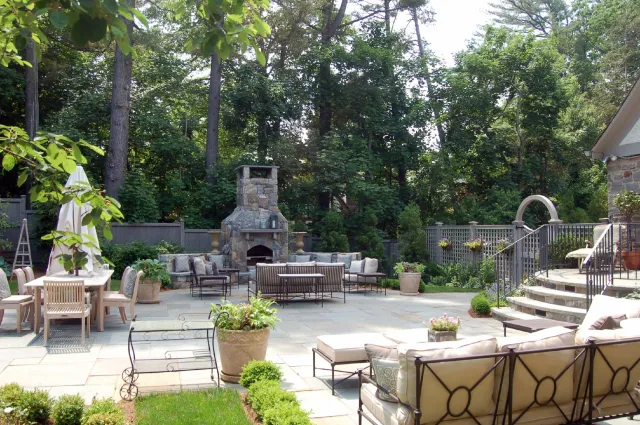 Backyard patio with stone fireplace, outdoor furniture, potted plants, and tall trees in the background.